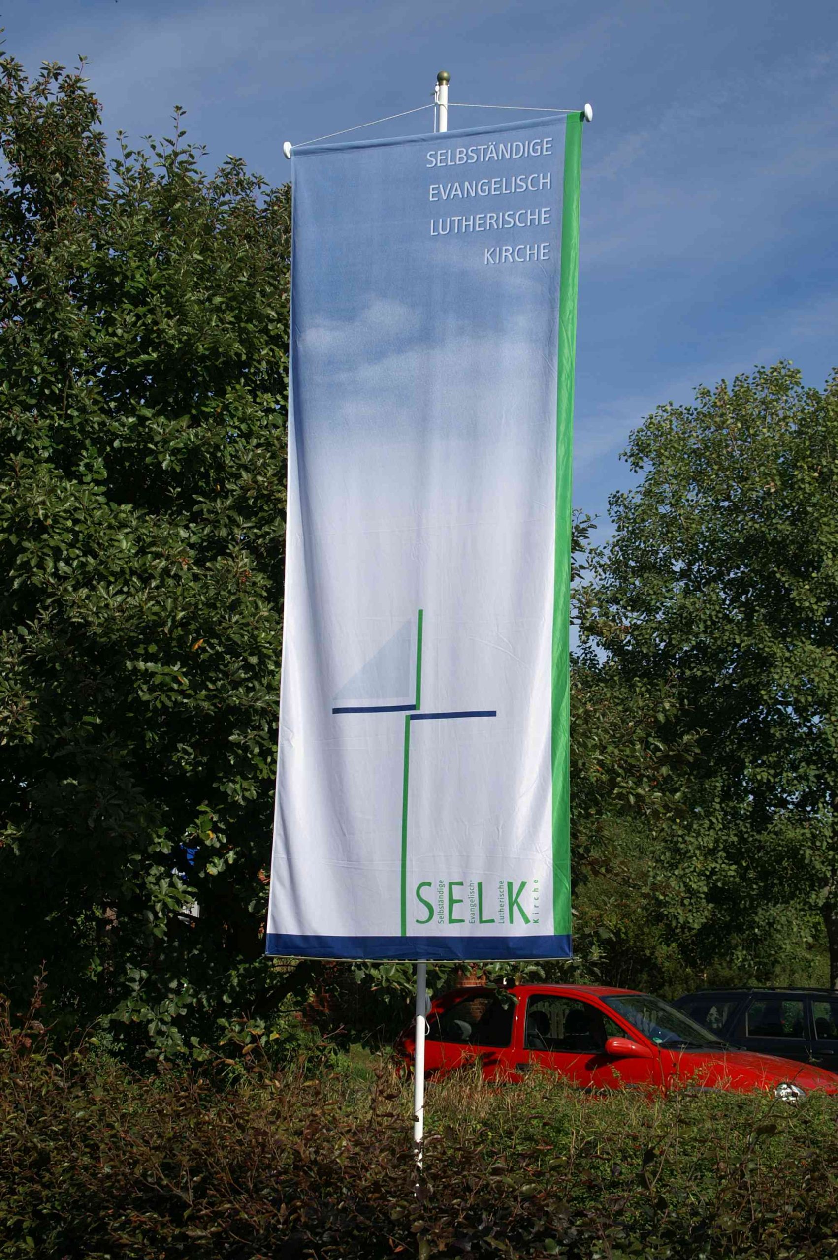 Fahne mit Logo der Selbst&auml;ndigen Evangelisch-Lutherischen Kirche vor dem Parkplatz der Holzkirche